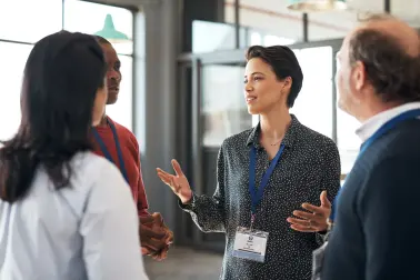 Kollegen stehen in einem Büro zusammen. Eine Frau spricht und gestikuliert, während die anderen zuhören.
