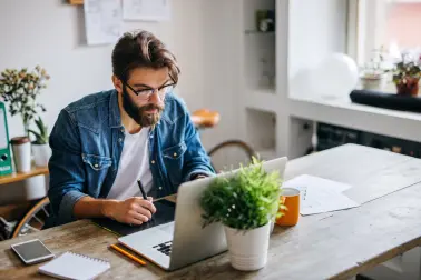 Mann mit Brille sitzt an einem Tisch und arbeitet mit Stift und Tablet vor einem Laptop. Auf dem Tisch steht eine Pflanze.