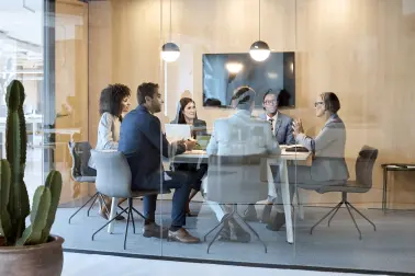 Sechs Personen sitzen in einem Besprechungsraum. Sie sind durch Glas zu sehen.