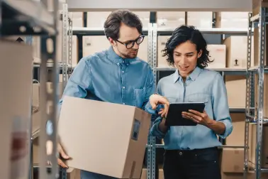 Frau steht in einer Lagerhalle mit vielen Kartons in Regalen. Sie schaut auf einem Tablet. Neben ihr steht ein Mann und trägt eine Kiste. Er zeigt auf etwas auf dem Tablet. 