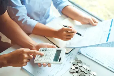 Zwei Personen sitzen an einem Schreibtisch. Vor ihnen liegen viele Dokumente, Rechnungen und Münzen. Eine Person benutzt einen Taschenrechner. 