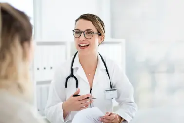 Ärztin in weißem Kittel und Stethoskop um den Hals sitzt an einem Tisch. Vor ihr liegen Unterlagen und sie hält einen Stift in der Hand. Ihr gegenüber sitzt eine Patientin.