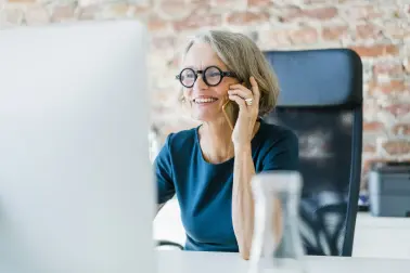 Ältere Frau sitzt an einem Schreibtisch. Sie schaut auf einen Bildschirm und telefoniert. Sie lächelt.