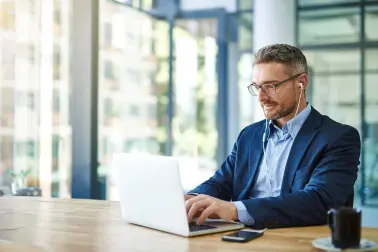 Mann in Anzug sitzt an einem Tisch. Er schreibt etwas auf seinem Laptop. Er hat weiße Kabelkopfhörer in den Ohren, welche in sein Smartphone gesteckt sind. Das Smartphone liegt neben dem Laptop auf dem Tisch.